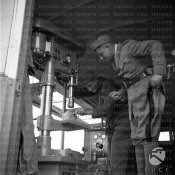 3_Un carpentiere del genio usa un trapano verticale all'interno di un'auto-officina osservato.jpg